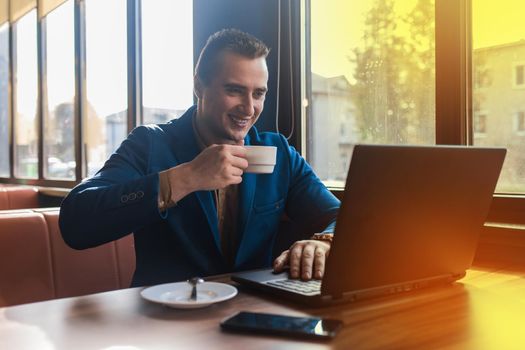 A business smiling positive man businessman a stylish portrait of Caucasian appearance in a jacket, works in a laptop or computer, drink coffee on coffee break, sitting at a table by the window.