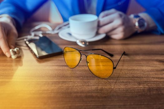 Sunglasses, mobile phone or smartphone and a mug of coffee on the table in a cafe with the hand of a businessman.