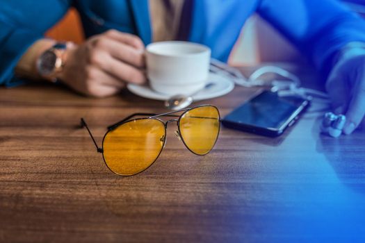 Sunglasses, mobile phone or smartphone and a mug of coffee on the table in a cafe with the hand of a businessman.