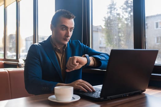 A business surprised man businessman a stylish of Caucasian appearance in a jacket, works in a laptop or computer, sitting at a table by the window in a cafe and looks at the time on a wrist watch.