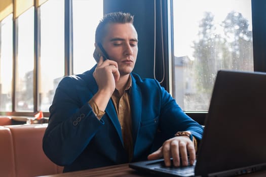 A business man stylish serious businessman in an attractive European-looking suit works in a laptop, talks on a cell or mobile phone, sitting at a table in a cafe by the window.