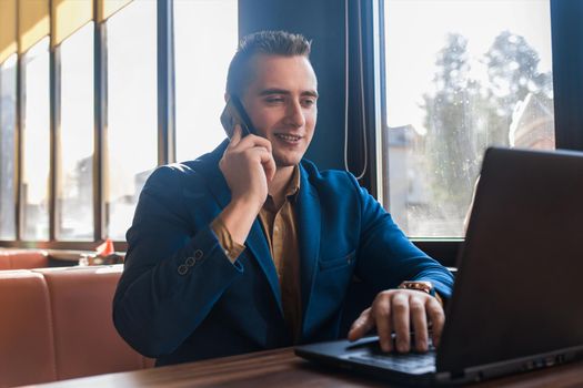 A business man stylish positive smiling businessman in an attractive European-looking suit works in a laptop, talks on a cell or mobile phone, sitting at a table in a cafe by the window.