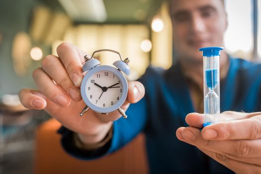 The hand of a male businessman holds a clock alarm time and an hourglass, close-up.