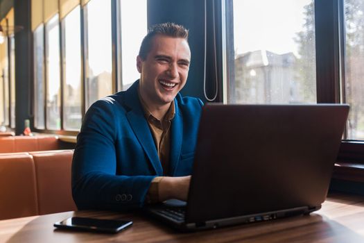 A business man happy positive businessman a stylish portrait of Caucasian appearance in a jacket works in a laptop or computer, sitting at a table by the window in a cafe.