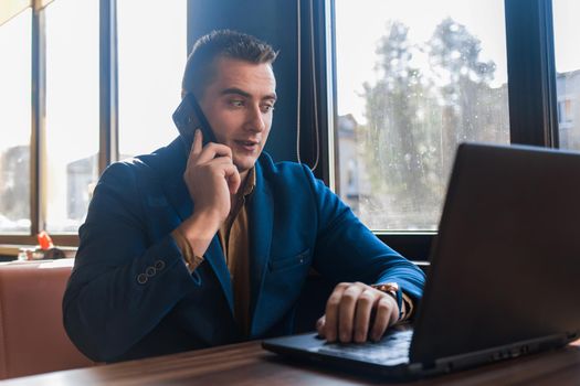 A business man surprised businessman in an attractive European-looking suit works in a laptop, talks on a cell or mobile phone, sitting at a table in a cafe by the window.