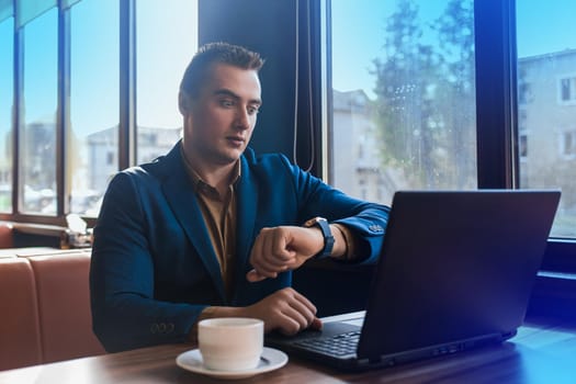 A business surprised man businessman a stylish of Caucasian appearance in a jacket, works in a laptop or computer, sitting at a table by the window in a cafe and looks at the time on a wrist watch.