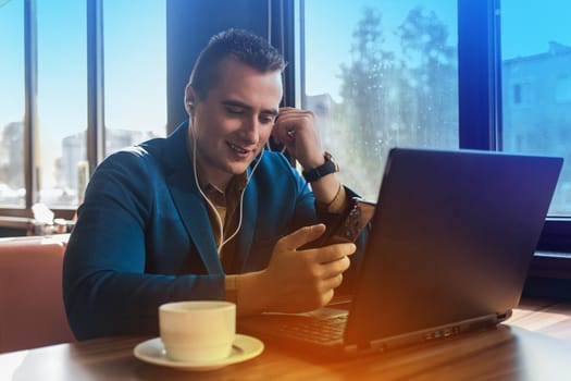 A business smiling man stylish businessman in an attractive European-looking suit works in a laptop, listens to music with headphones and drinks coffee sitting at a table in a cafe by the window.