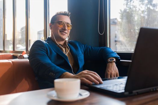 A business positive man smiling businessman a stylish of Caucasian appearance, in sunglasses, works in a laptop or computer, sitting at a table by the window in a cafe and listening to music.