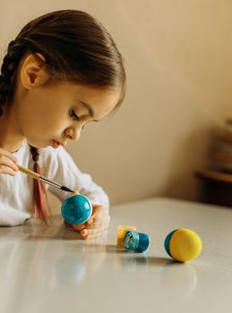 Portrait of a Ukrainian girl. the child paints the egg blue and yellow. The child loves and supports his country. Cute little child girl painting with blue and yellow colors Easter eggs. Hands of a girl with a easter egg. Close-up.