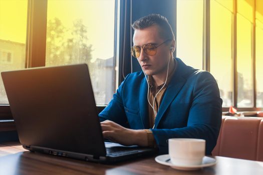 A business man stylish serious businessman in an attractive European-looking suit works in a laptop, listens to music with headphones and drinks coffee sitting at a table in a cafe by the window.