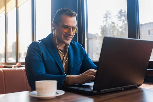 A business man stylish smiling businessman in an attractive European-looking suit works in a laptop, listens to music with headphones and drinks coffee sitting at a table in a cafe by the window.