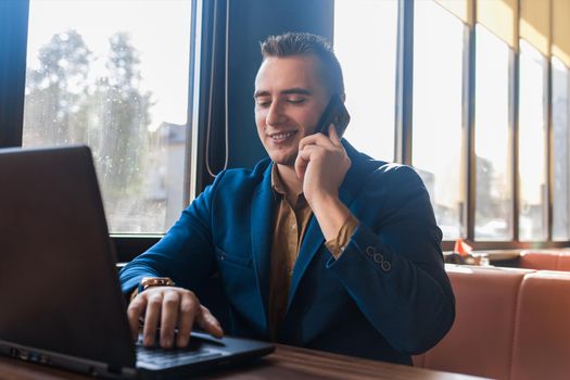 A business man stylish positive smiling businessman in an attractive European-looking suit works in a laptop, talks on a cell or mobile phone, sitting at a table in a cafe by the window.