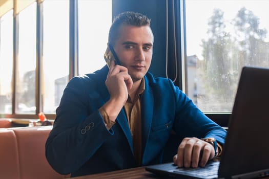 A business man surprised businessman in an attractive European-looking suit works in a laptop, talks on a cell or mobile phone, sitting at a table in a cafe by the window.