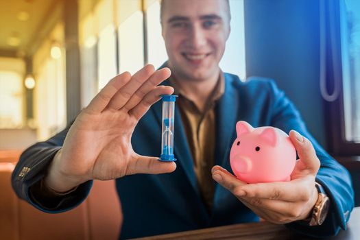 The hands of a smiling businessman in a suit hold an hourglass of time and a piggy bank of savings.