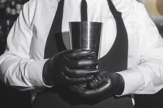 The hands of a professional bartender man in black latex gloves holds a tool for mixing and making shaker cocktails.