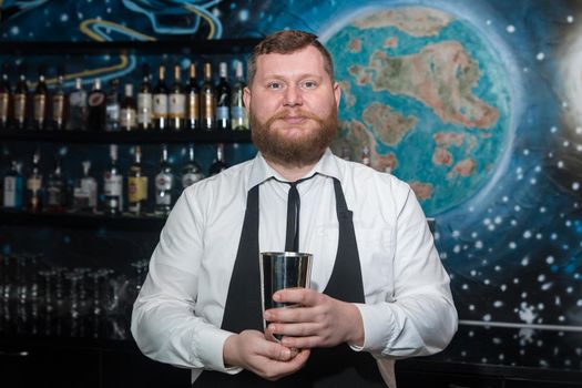 A bearded adult man of European appearance, a professional bartender, holds in his hands a tool for preparing and mixing alcoholic cocktails in a nightclub.