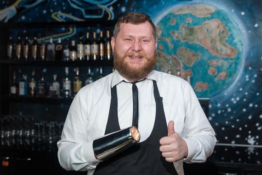 A bearded smiling funny adult man of European appearance, a professional bartender, holds in his hands a tool for preparing and mixing alcoholic cocktails in a nightclub.