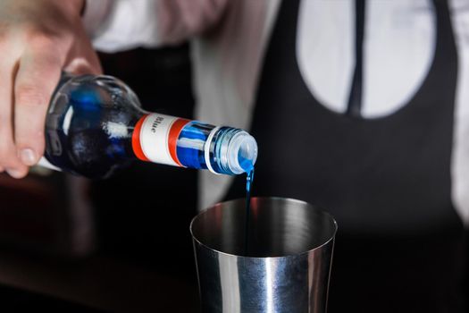 The hand of a professional bartender pours blue syrup into a tool for preparing and mixing alcoholic cocktails shaker, close-up.