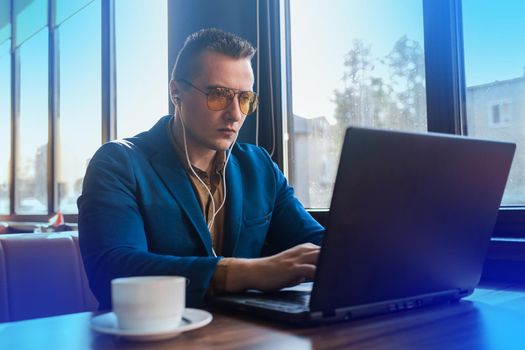 A business man stylish serious businessman in an attractive European-looking suit works in a laptop, listens to music with headphones and drinks coffee sitting at a table in a cafe by the window.