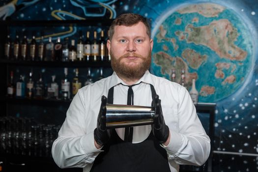 A bearded adult man of European appearance, a professional bartender, holds in his hands in black latex gloves a tool for preparing and mixing alcoholic cocktails in a nightclub.