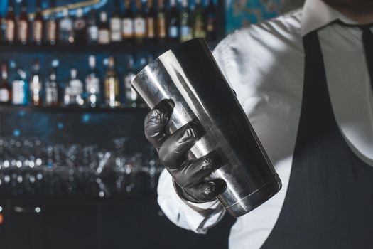 The hand of a professional bartender in a black latex glove with a bar instrument for mixing and preparing alcoholic cocktails with a shaker.