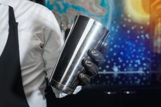 The hand of a professional bartender in a black latex glove with a bar instrument for mixing and preparing alcoholic cocktails with a shaker.