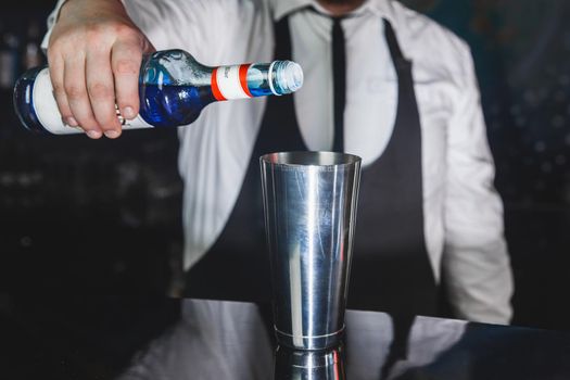 The hand of a professional bartender pours blue syrup into a tool for preparing and mixing alcoholic cocktails shaker, close-up.