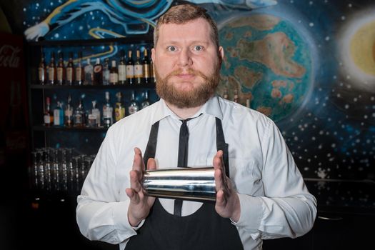 A bearded adult man of European appearance, a professional bartender, holds in his hands a tool for preparing and mixing alcoholic cocktails in a nightclub.