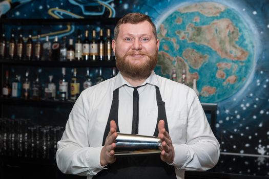 A bearded adult man of European appearance, a professional bartender, holds in his hands a tool for preparing and mixing alcoholic cocktails in a nightclub.