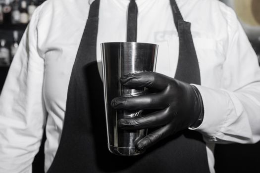 The hand of a professional bartender man in black latex glove holds a tool for mixing and making shaker cocktails.