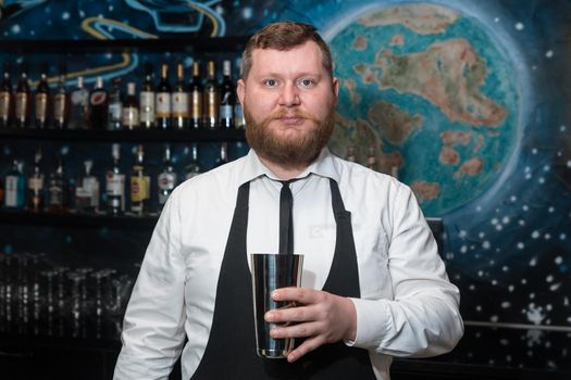 A bearded adult man of European appearance, a professional bartender, holds in his hands a tool for preparing and mixing alcoholic cocktails in a nightclub.