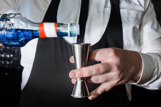 The hand of a professional bartender pours blue syrup into a tool to control the ingredients added to a jigger cocktail or measuring cup. The process of preparing a cocktail.