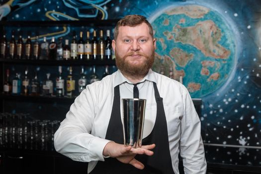 A bearded adult man of European appearance, a professional bartender, holds in his hands a tool for preparing and mixing alcoholic cocktails in a nightclub.