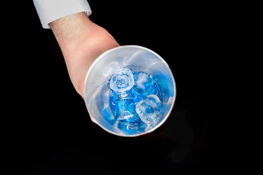 The hand of professional bartender takes a tool for mixing and making alcoholic cocktails shaker with blue cold syrup with ice in on a black background, close-up.