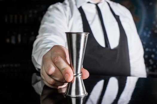 The hand of a professional bartender holds a jigger tool or measuring cup to control the ingredients added to the cocktail.