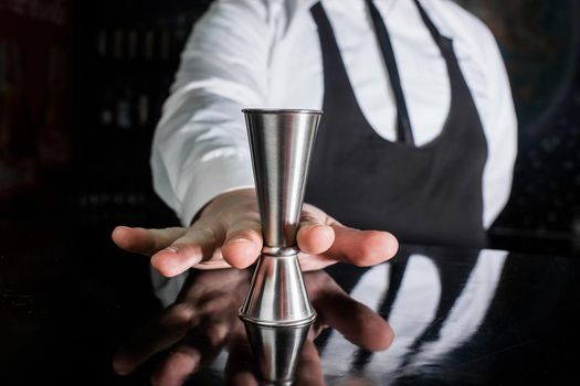 The hand of a professional bartender holds a jigger tool or measuring cup to control the ingredients added to the cocktail.