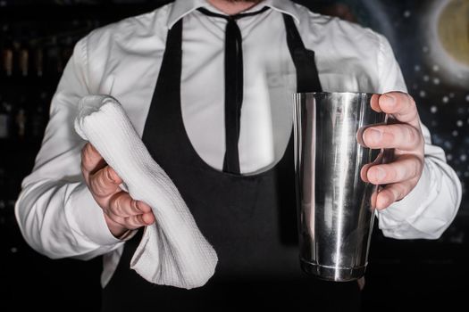 A professional bartender holds a metal shaker tool for mixing and making alcoholic cocktails and wipes it with a white cloth.