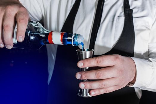 The hand of a professional bartender pours blue syrup into a tool to control the ingredients added to a jigger cocktail or measuring cup. The process of preparing a cocktail.