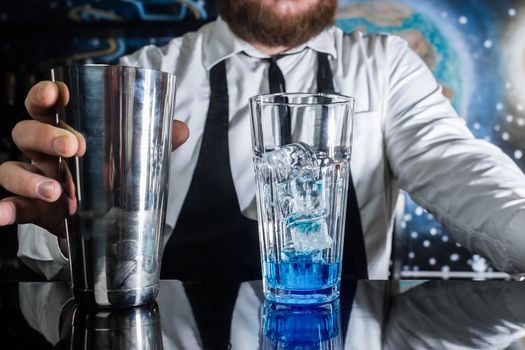 The process of preparing a cocktail. The hand of a professional bartender holds a metal shaker liquid mixing tool next to a glass with ice and blue syrup.