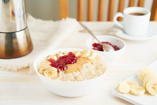 Oatmeal porridge, healthy vegan diet breakfast with strawberry jam, peanut butter, banana, chia on white wooden light background. Side view