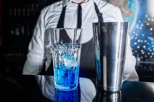 A glass with ice and blue syrup stands on the bar counter against the background of a professional bartender, next to a tool for mixing and preparing alcoholic cocktails with a metal shaker.