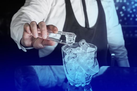 The hand of a professional bartender holds plastic ice cube tongs next to a cold ice dish.