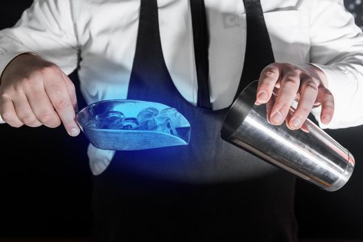 The hands of a professional bartender add ice in a scaple to a tool for preparing and mixing alcoholic cocktails, a metal shaker.