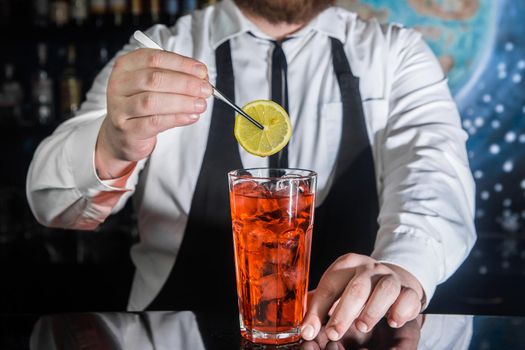 A professional bartender puts a slice of lemon in a red chilled alcoholic cocktail with bar tweezers in glass at a nightclub counter.