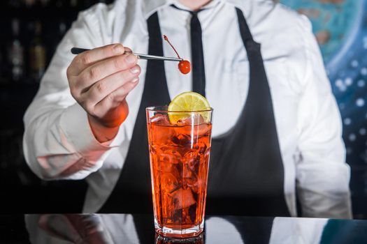 A professional bartender puts a cherry in a red chilled alcoholic cocktail with bar tweezers in glass at a nightclub counter.