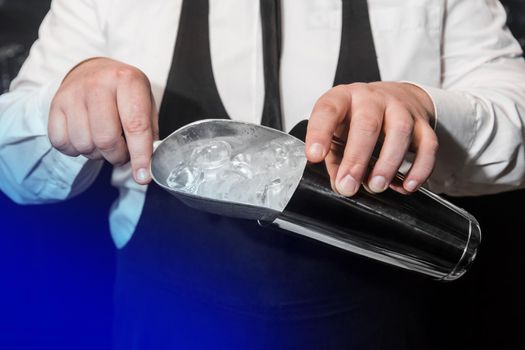 The hands of a professional bartender add ice in a scaple to a tool for preparing and mixing alcoholic cocktails, a metal shaker.