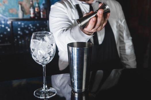 The hands of a professional bartender pour syrup into a measuring glass of jigger in a metal tool for preparing and stirring alcoholic cocktails of shaker drinks, and glasses with ice cubes.