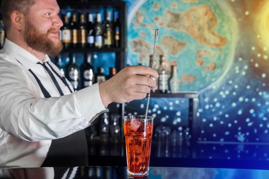 Adult bearded smiling bartender is professional and positive interferes with a bar spoon red alcoholic cocktail drink in glass on the bar counter. Serving a cocktail.