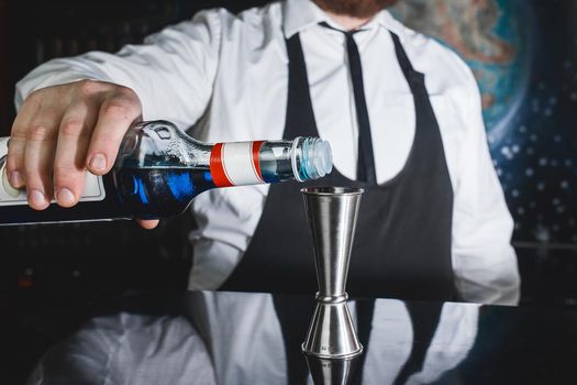 The hand of a professional bartender pours blue syrup into a tool to control the ingredients added to a jigger cocktail or measuring cup. The process of preparing a cocktail.