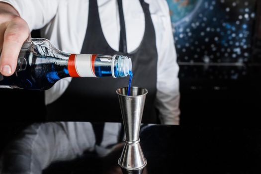 The hand of a professional bartender pours blue syrup into a tool to control the ingredients added to a jigger cocktail or measuring cup. The process of preparing a cocktail.